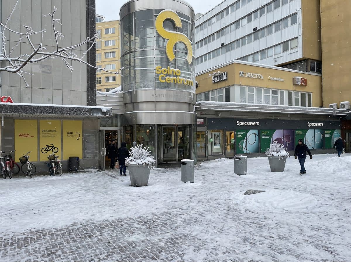 Bilden visar entréområdet till Solna Centrum från utsidan under vintertid med snö på marken och byggnaderna. En stor, rundad glasstruktur med en gul logotyp märkt "Solna Centrum" fungerar som huvudentré. Till vänster om entrén finns en gul vägg med s