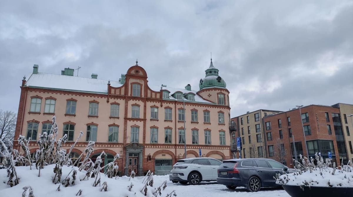 Bilden visar en gatuvy under dagtid med snötäckt mark. I förgrunden syns snö, buskar och växter täckta med frost och snö. I mitten av bilden dominerar en äldre byggnad med ljusrosa fasad, rött tegeldekoration och ett grönt kupoltak. Byggnaden har mån
