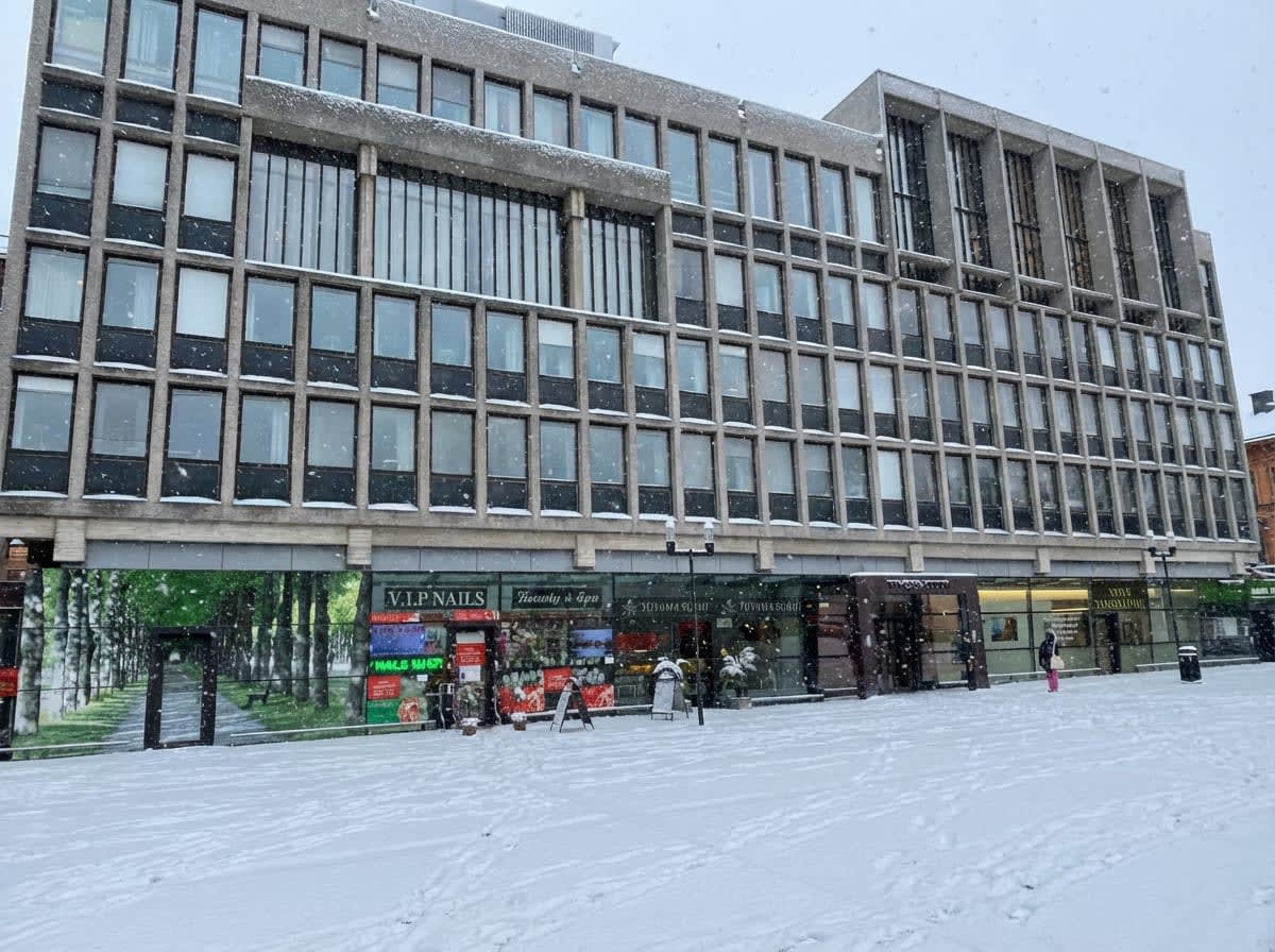 Bilden visar ett stort, flervåningshus under ett snöfall. Husets fasad består av betong och har ett stort antal rektangulära fönster arrangerade i ett rutmönster. Delar av fönsterkarmarna och horisontella avsatser är täckta med snö. På gatunivå finns