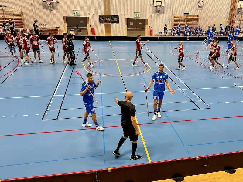 Bilden visar en inomhusarena med en blå plan, utmärkt med vita, röda och gula linjer, där en innebandymatch spelas. På planen befinner sig flera spelare klädda i antingen blåa eller röda uniformer, samt två domare i svarta kläder. En av domarna pekar