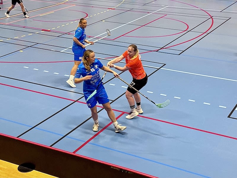 En bild tagen inomhus som visar en pågående innebandymatch. Tre kvinnliga spelare är synliga, varav två är klädda i blåa dräkter och en i en orange överdel med svarta shorts. Spelarna är engagerade i spelet på en blå innebandyplan med svarta, vita oc