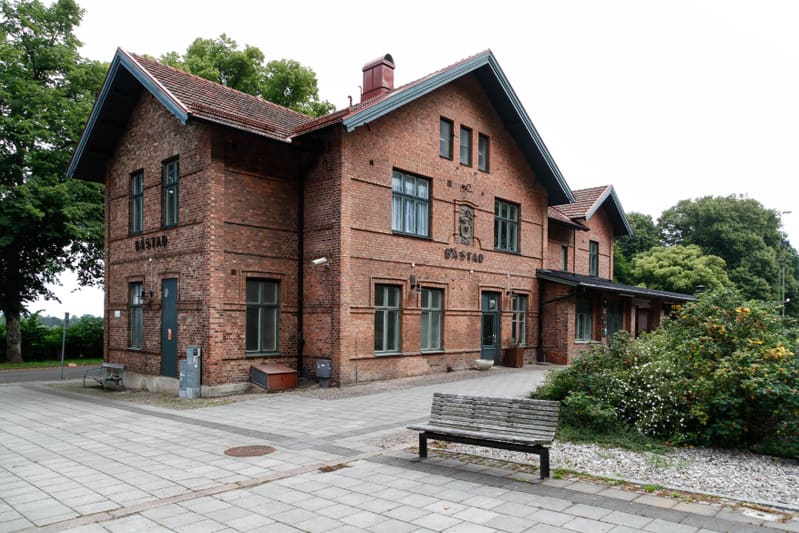 Bilden visar en tegelbyggnad, troligen en stationsbyggnad, sedd från en lätt upphöjd vinkel. Byggnaden har ett tegeltak med takpannor och flera fönster med gröna karmar. Ovanför några fönsterpartier syns texten "BÅSTAD". Framför byggnaden finns ett s