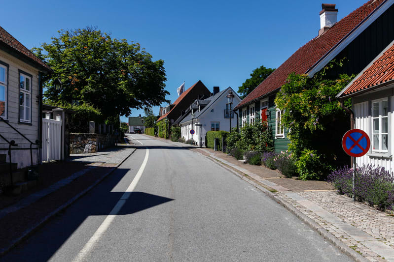 En bild tagen från gatan av en villagata med flera hus i olika färger och byggstilar längs sidorna. Till vänster i förgrunden syns skuggor över trottoaren och gatan. Gatan har en vit heldragen linje i mitten. Husen är omgivna av grönska, buskar och t