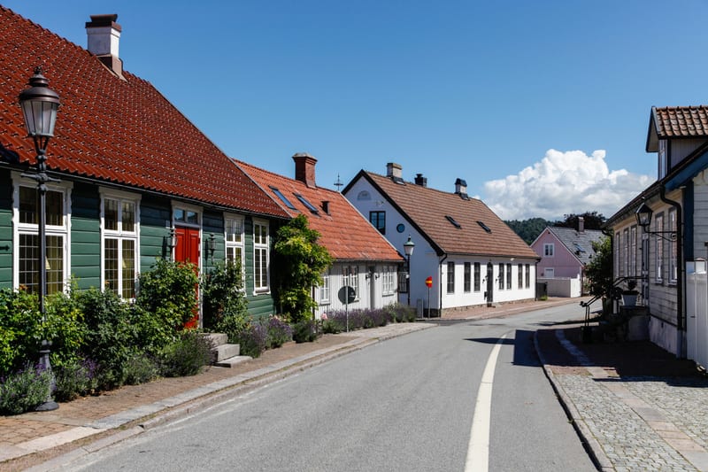 Bilden visar en gata i en stad eller by under dagtid med flera hus längs sidorna. På vänster sida syns ett grönmålat trähus med ett rött tak och vita fönsterkarmar. En röd dörr är synlig i mitten av fasaden. Framför detta hus finns buskar och planter