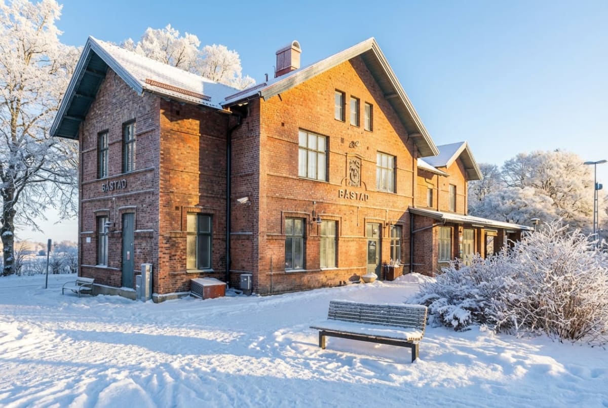 En bild tagen utomhus under dagtid som visar en tegelbyggnad med texten "BÅSTAD" på fasaden, upplyst av solljus från höger. Byggnaden har flera fönster och en skorsten. Runt byggnaden ligger ett snötäckt landskap med spår i snön. Till vänster i bilde