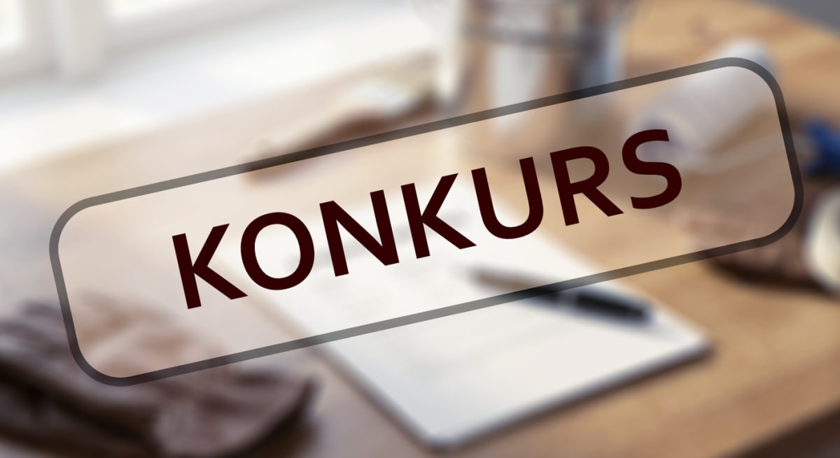 Bilden visar ett skrivbord eller en bordsyta med suddiga objekt i bakgrunden och förgrunden. I mitten av bilden finns en rektangulär, halvtransparent ruta med rundade hörn som innehåller texten "KONKURS" i stora, mörkbruna bokstäver. Ytan på bordet verkar vara av trä i en ljusbrun nyans. I den suddiga bakgrunden kan man vagt urskilja vad som ser ut som fönster eller en ljus vägg. Bland de otydliga objekten syns en metallbehållare, möjligen en mugg eller hink, en liten anteckningsbok eller block samt en penna. Också otydliga tyger eller handskar kan skönjas i det nedre vänstra hörnet. Den övergripande bilden har ett dämpat, lätt oskarp fokus med texten i förgrunden som det skarpaste elementet.
