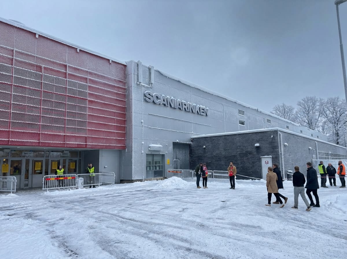 En utomhusbild tagen under dagtid föreställer framsidan av en stor byggnad med texten "SCANIARINKEN" på fasaden. Byggnadens vänstra sida har en röd gallerstruktur, medan resten av fasaden är grå. Taket och delar av byggnaden är täckta med snö och is. Entrén till byggnaden är synlig till vänster, och vita avspärrningsstaket med röda och gula detaljer leder fram till den. Flera personer, både enskilda och i små grupper, befinner sig på den snötäckta marken framför byggnaden. Några av personerna vid entrén bär gula reflexvästar. Marken består av snö och is med spår av gångtrafik. I bakgrunden syns snötäckta träd och en gråmulen himmel.