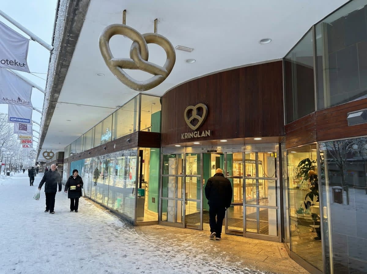 Bilden visar entréområdet till ett köpcentrum eller en galleria som heter Kringlan. Ingången har stora glasdörrar och ovanför dörren sitter en gyllene logotyp som föreställer en kringla med ett hjärta i mitten, samt texten "KRINGLAN" under. Framför entrén går en person i mörk jacka mot dörrarna. Till vänster om entrén sträcker sig en gångväg täckt med snö där flera personer promenerar. Byggnaden har stora skyltfönster längs gångvägen och ovanför dem hänger flera vita flaggor eller banderoller med text, bland annat "apteket" och "ICA". Takutsprånget ovanför butikerna är täckt av ett tunt lager snö. Markytan är täckt av snö och is. Himlen är ljus och molnig.