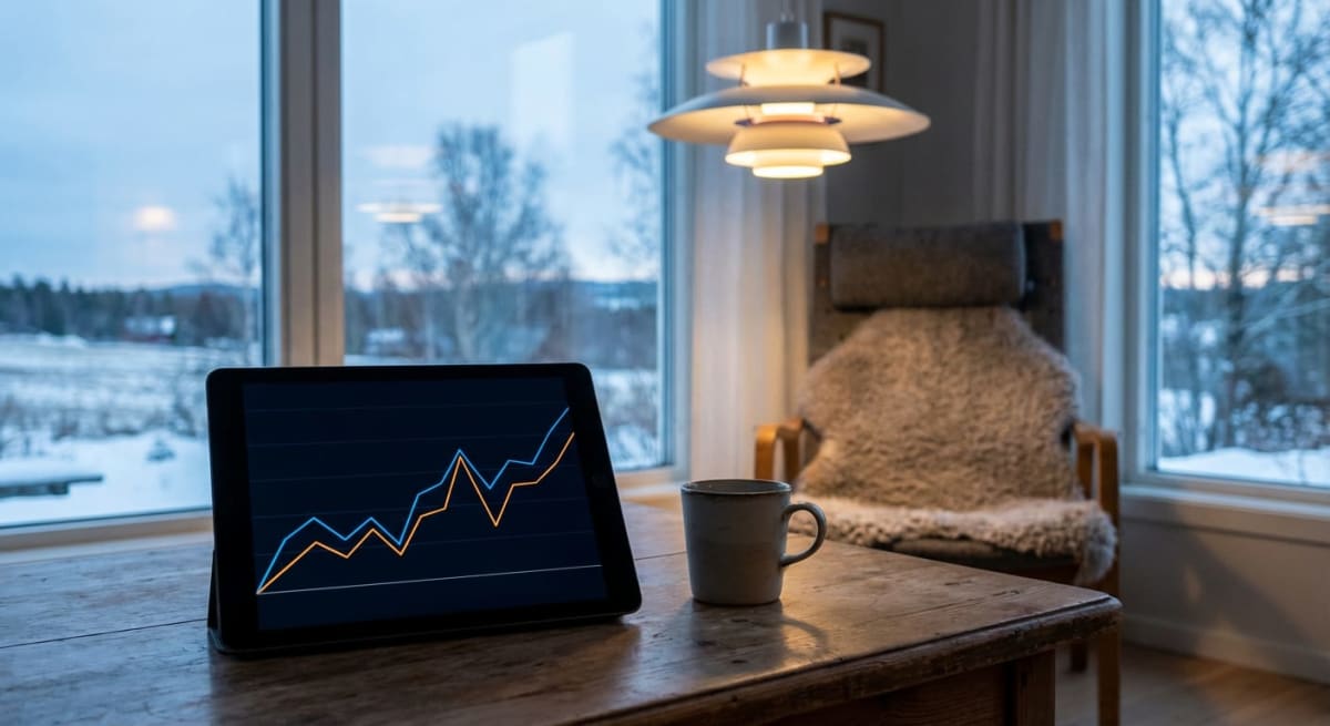 Bilden visar ett landskap täckt av snö sett genom ett stort fönster. I förgrunden syns ett träbord med en surfplatta som visar ett diagram med två linjer, en blå och en orange, mot en mörk bakgrund. Bredvid surfplattan står en grå mugg. Till höger om bordet finns en karmstol klädd med ett lurvigt fårskinn. Ovanför stolen hänger en vit taklampa med flera skärmar. Utanför fönstret är det ett vinterlandskap med träd och snö. Ljuset indikerar troligen skymning eller gryning, eller en mulen vinterdag.