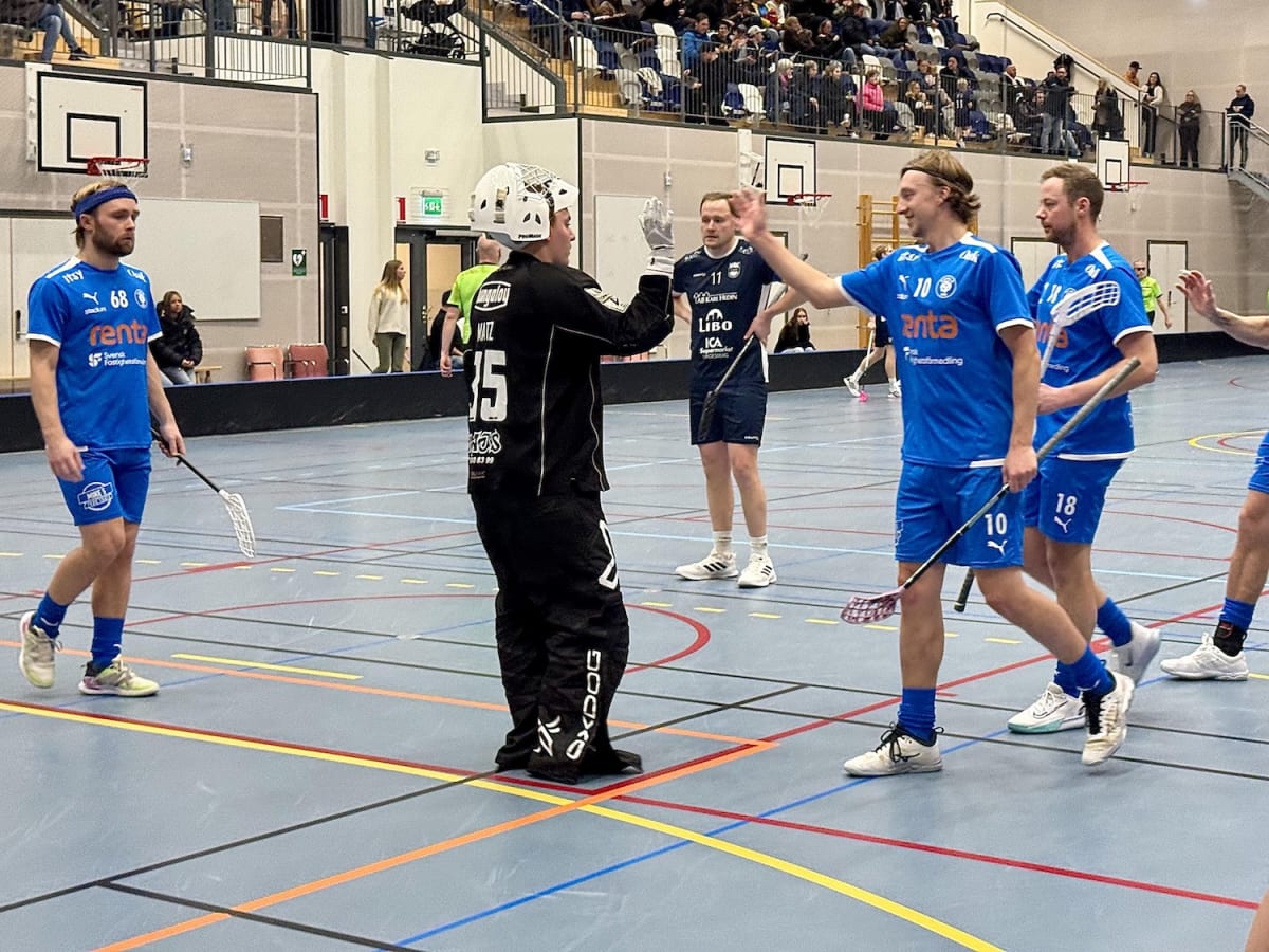 Bilden visar en inomhusidrottsanläggning där flera personer spelar eller ser på innebandy. I förgrunden syns tre spelare i blåa dräkter och en målvakt i svart dräkt. Målvakten har en vit hjälm och stor skyddsutrustning. Två av de blåklädda spelarna h