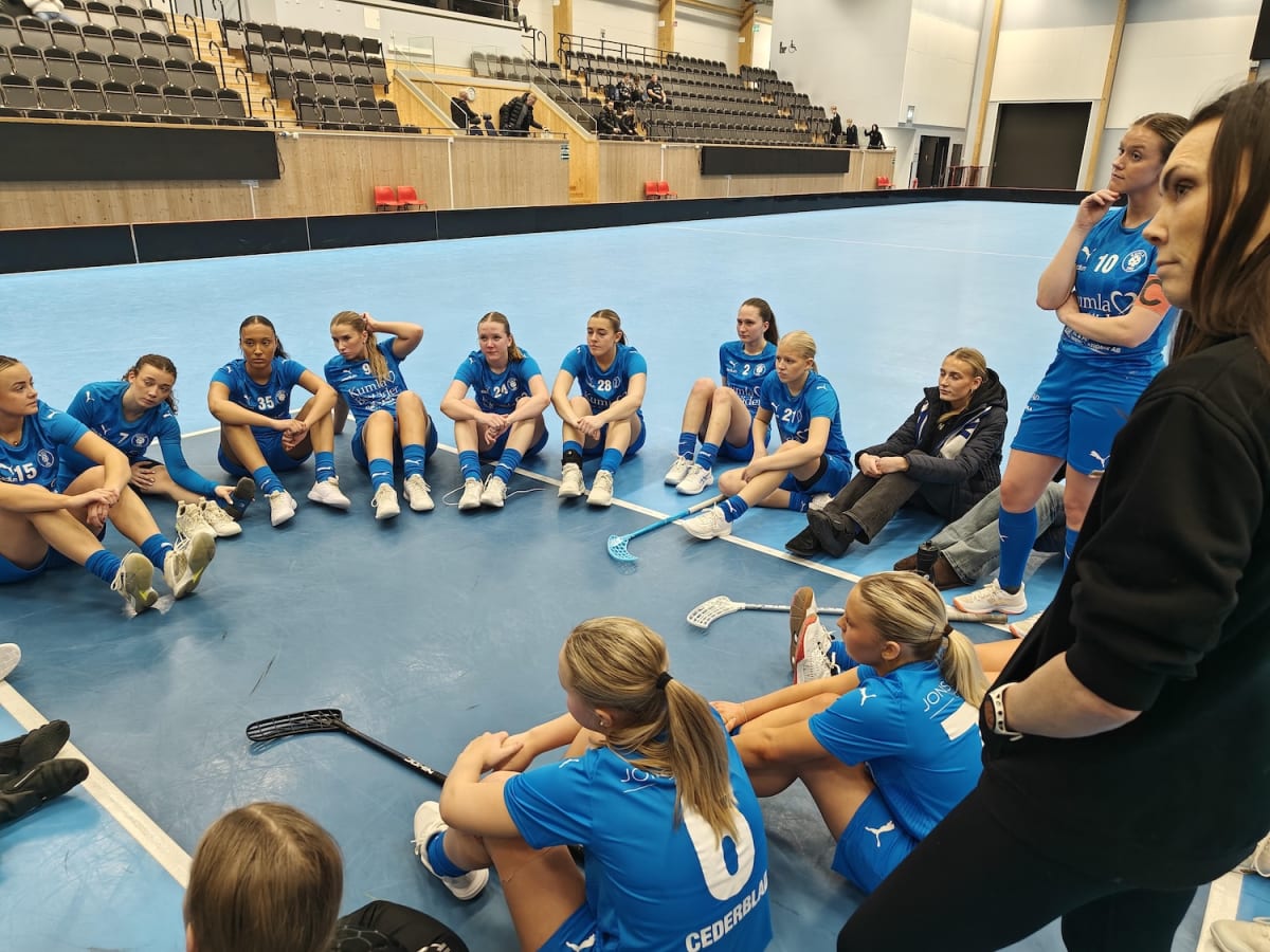 En grupp kvinnor är samlade på en blå idrottshall som liknar en innebandyplan. De flesta kvinnorna sitter ner i en halvcirkel, några är klädda i blå matchställ och andra i fritidskläder. På matchställen syns siffror som 15, 7, 35, 9, 24, 28, 2, och 1