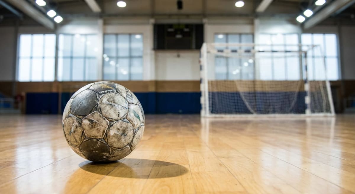 En nedifrån-och-upp-vy i ögonhöjd av en sliten, smutsig fotboll som ligger på ett glänsande trägolv i en idrottshall. Bollen är i förgrunden och består av gråvita och mörkare paneler. I bakgrunden syns en vit målställning med ett blått nät och väggar med blå och beigea fält under stora fönster som släpper in ljus. Taket visar vita lysrörsbelysningar. Golvet reflekterar ljuset från belysningen och fönstren.