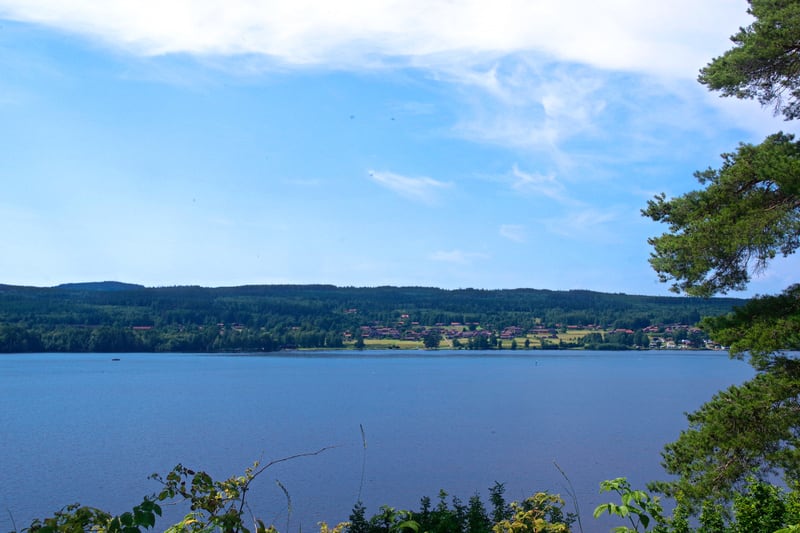 Bilden visar ett landskap med en stor sjö i förgrunden och ett skogsbeklätt land på andra sidan. Vid horisonten, bortom sjön, syns en rad hus med mörka tak, omgivna av grönska, vilket indikerar en by eller mindre bosättning. Himlen är ljusblå med spr