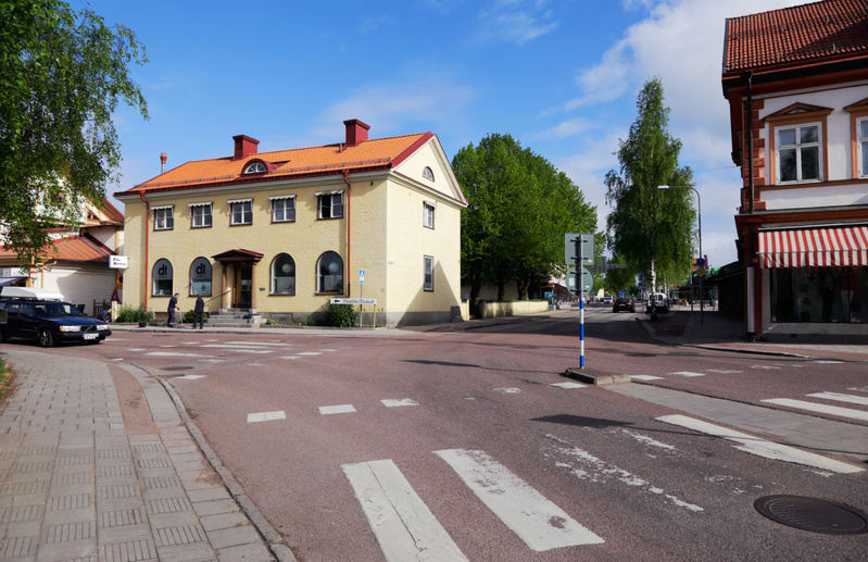 Bilden visar ett gathörn i en stadsmiljö under dagtid. I förgrunden syns en rödbrun asfaltväg med vita övergångsställen. Till vänster finns en trottoar med plattor och kantsten. En svart personbil ses delvis på vägen till vänster. En gul byggnad med