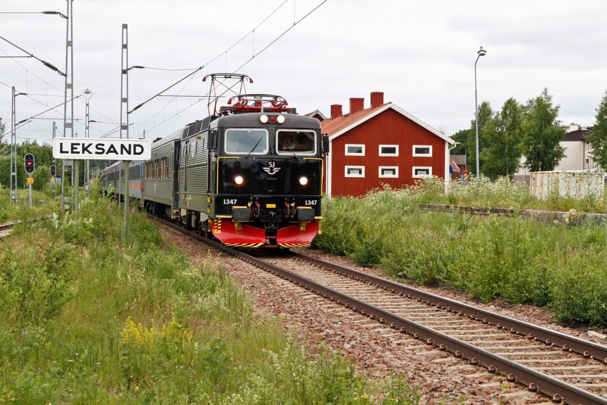 En svart SJ-lok, nummer 1347, syns på ett järnvägsspår i förgrunden, med den främre delen av tåget mot betraktaren. Tåget har röda detaljer på fronten och flera vagnar bakom loket. Till vänster om tåget står en vit skylt med texten "LEKSAND" i svart