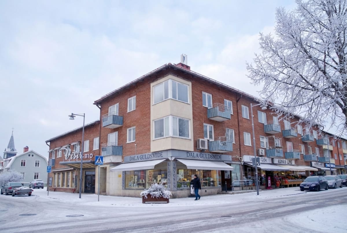 Bilden visar en snötäckt stadsgata under dagtid. I förgrunden till höger syns ett snötäckt träd med bara grenar. Till vänster i bilden, över gatan, står en stor tegelbyggnad med flera balkonger och butikslokaler på bottenvåningen. En av butikerna har en skylt som lyder "DALA GULDSMIDE". Utanför denna butik står en person klädd i mörk jacka och byxor. Framför byggnaden finns en snötäckt blomlåda. På gatan och trottoarerna ligger det snö, men vägbanan är plogad. Några bilar, både parkerade och körande, syns på gatan. I bakgrunden till vänster skymtar ett hus med en kyrktornsliknande byggnad bakom. Himlen är ljus och molnig.