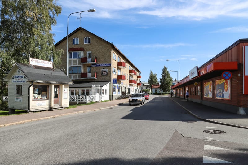 Bilden visar en gatuscen i en småstad under dagtid med klarblå himmel. Till vänster i förgrunden syns en liten vit korvkiosk med skylten "Vild-Hasses Korvböd" och "Renkorv Vild-Hasses Fjällprodukter". Bakom kiosken står ett flervåningshus i tegel med