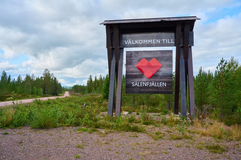 Bilden visar en välkomstskylt vid en grusväg i ett skogslandskap. Skylten, som står på höger sida av grusvägen, är byggd av mörkt trä och har en vit text som lyder "VÄLKOMMEN TILL" följt av en röd hjärtformad logotyp och texten "SÄLENFJÄLLEN". Bakom