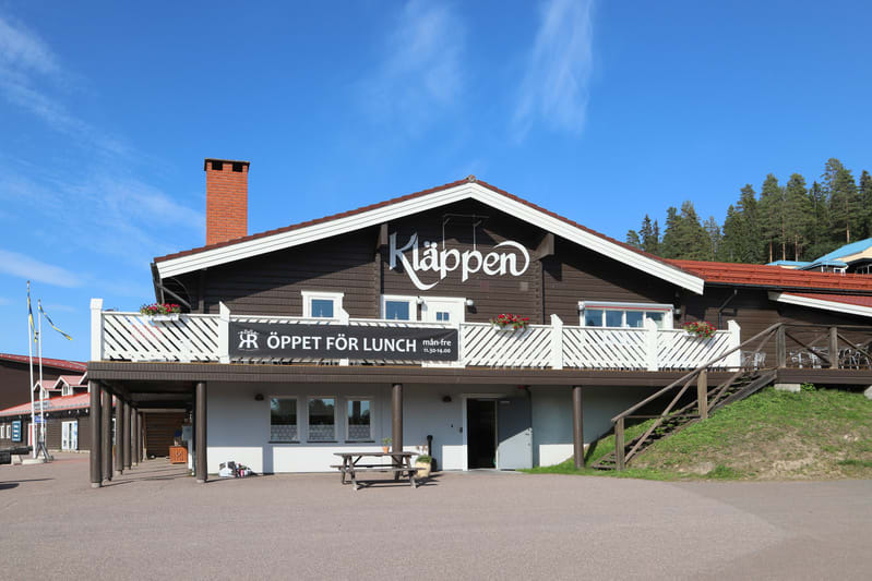 Bilden visar en stor byggnad med mörkbrun träpanel och vita detaljer under en klarblå himmel. På byggnadens framsida finns fönster, en balkong med vitt räcke och blomlådor med röda blommor. Ovanför balkongen står ordet "Kläppen" i stora vita bokstäve