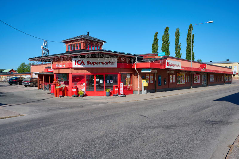 En stor, röd ICA Supermarket-byggnad med ett anslutande träfärgat område med svarta tak. Byggnaden har flera fönster och dörrar samt reklam för "ICA Supermarket". Framför entrén står en stor röd hästliknande staty med en figur i röda kläder bakom. På