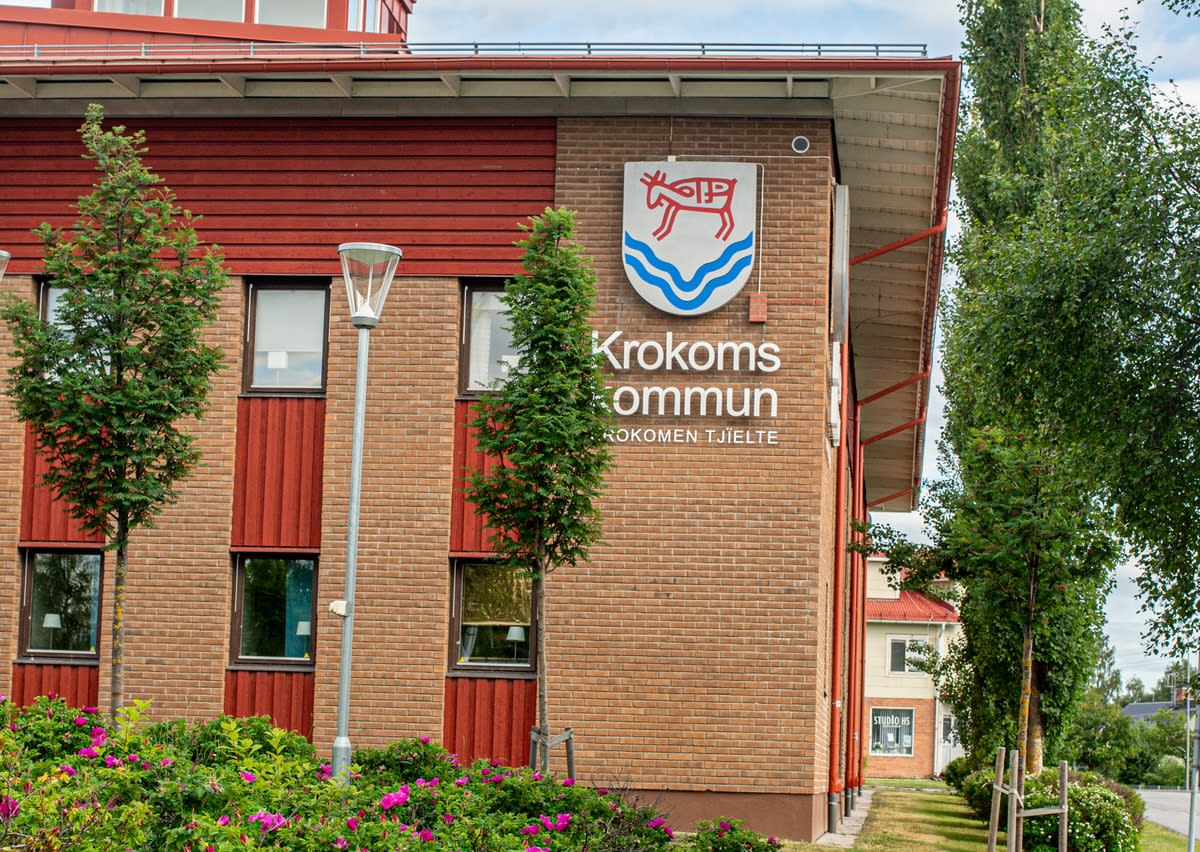 Bilden visar fasaden på en byggnad med tegelväggar och röda träpaneler runt fönstren. På tegelväggen sitter en skylt med Krokoms kommuns vapen, föreställande en röd älg och blå vågor, och texten "Krokoms kommun" på svenska och "KROKOMEN TJÏELTE" på s