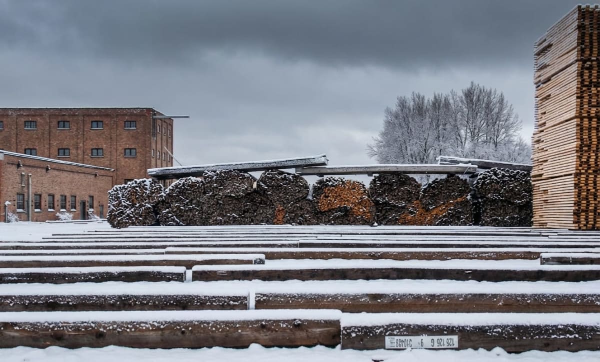 Bilden visar ett snöigt utomhusområde med flera strukturer och material. I förgrunden syns parallella rader av grova trästockar eller timmer som är täckta med ett lager snö. På en av stockarna i förgrunden finns en vit etikett med svart text. I mellangrunden syns stora högar av ved, delvis täckta med snö, placerade under ett tak eller skjul vars tak också är snötäckt. Bakom vedhögarna och till vänster i bilden reser sig en stor tegelbyggnad med många fönster och en mindre tegelbyggnad intill. Till höger om vedhögarna, utanför taket, står en hög stapel av plankor eller virke. I bakgrunden syns snötäckta, kala träd under en molnig grå himmel.