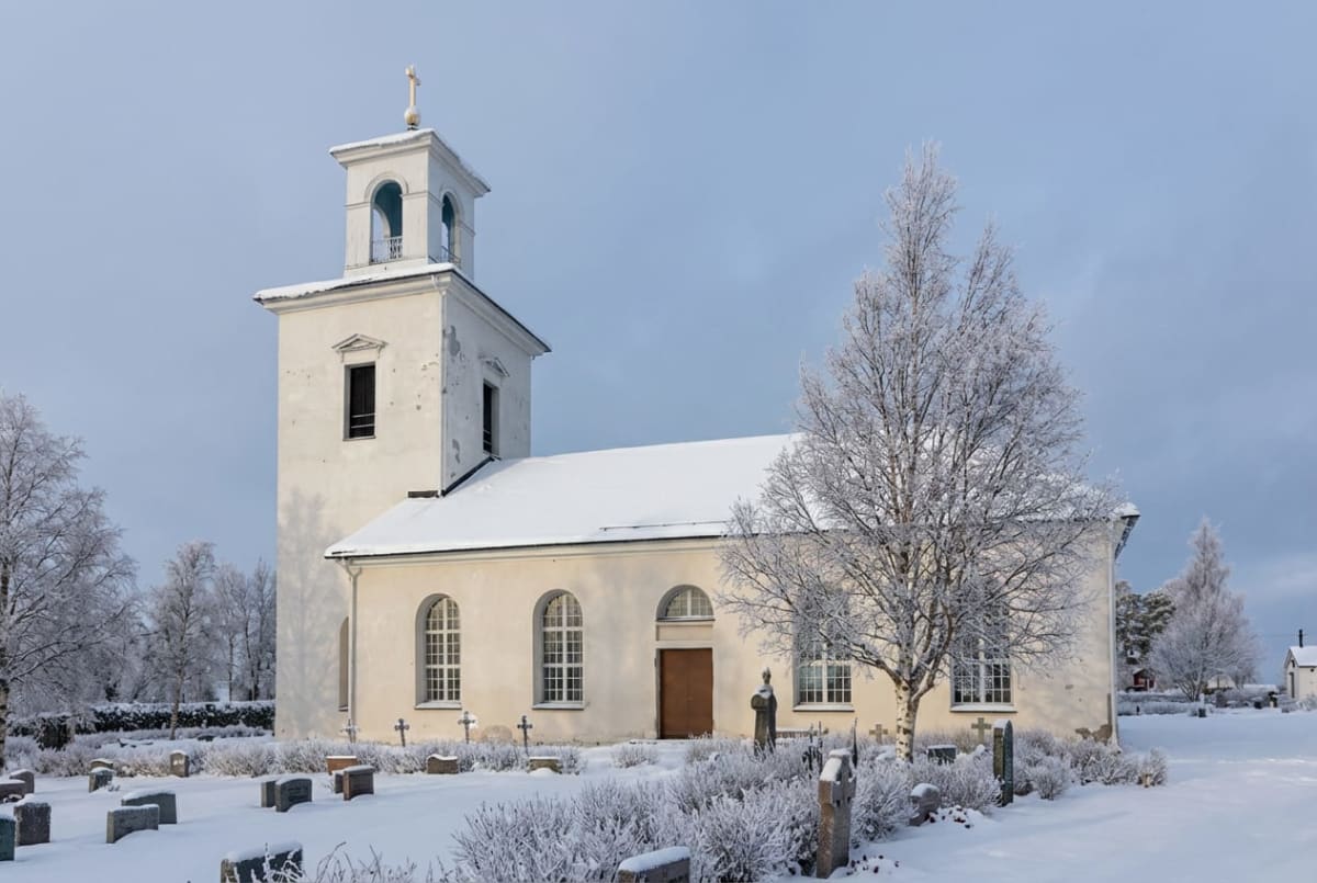 En bild som visar en vit kyrkbyggnad med ett torn och en kyrkogård täckt av snö. Kyrkbyggnaden har vita putsade väggar och ett mörkt tak täckt med ett lager snö. Tornet är också vitt, har en öppen klockvåning med valv och kröns av ett gyllene kors. På kyrkogården syns flera gravstenar som delvis är täckta av snö. Buskar och träd i förgrunden och runt om byggnaden är täckta med frost eller snö. Ett naket träd med förgrenade grenar står nära kyrkans framsida till höger. Himlen är ljusgrå och molnig. Bilden har ett lugnt och fridfullt intryck.