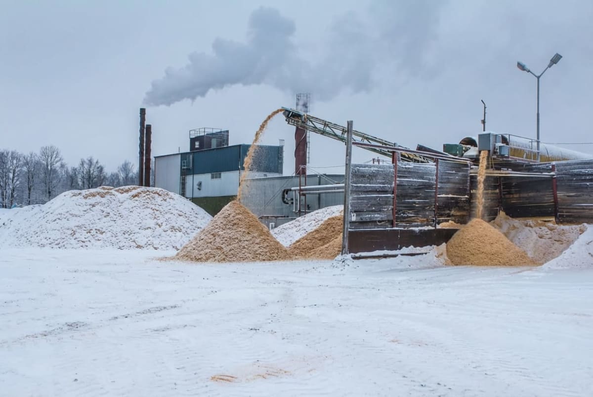 Bilden visar ett industriområde täckt av snö under en gråmulen dag. I förgrunden och mellan byggnaderna syns stora högar med snö. Olika sorters träflis eller sågspån matas via transportband och faller ner i flera separata högar. I bakgrunden syns flera industribyggnader, varav en har två höga skorstenar från vilka grå rök stiger upp mot himlen. En del av byggnaderna är klädda med blå panel. Till höger i bilden står en lampa med två riktade strålkastare som kastar ljus över området. Marken är täckt av ett vitt snötäcke med tydliga spår från fordon eller maskiner. Bara ett fåtal kala träd syns i bakgrunden till vänster.