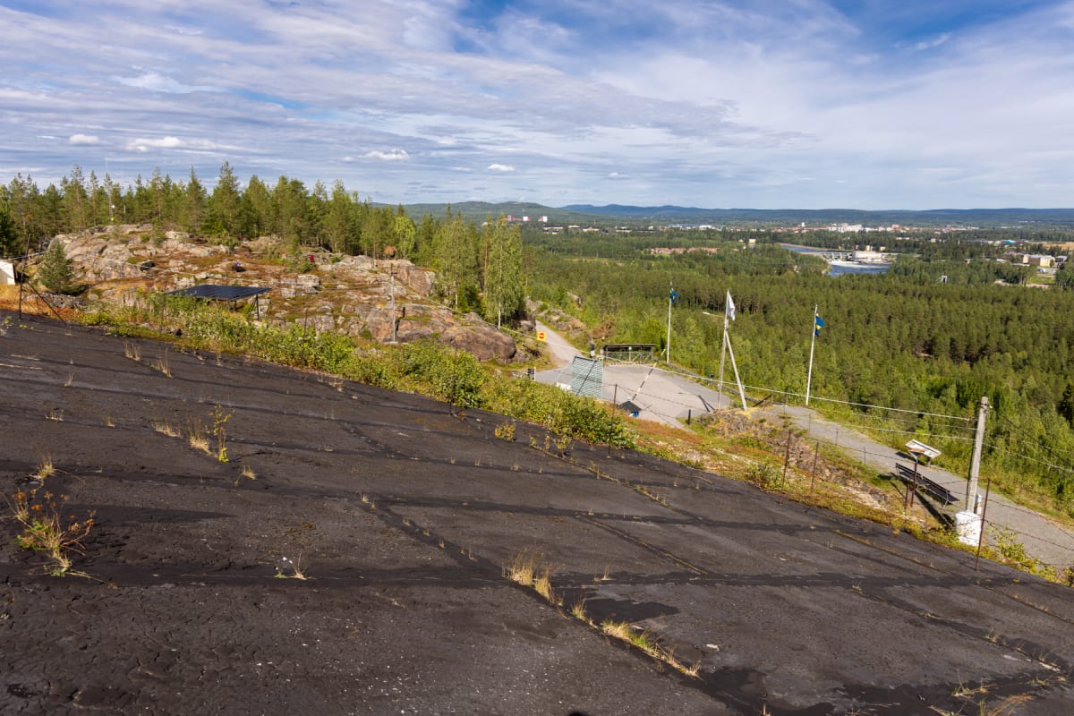Bilden visar ett landskap taget från en upphöjd position, med en svart, sluttande yta i förgrunden som är täckt med sprickor och sporadiskt växande gräs. I mitten av bilden finns ett bergigt område med träd och buskar, samt en mindre väg som slingrar