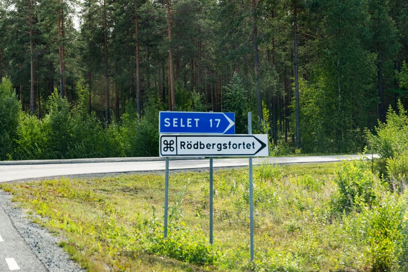 Bilden visar två vägskyltar som är monterade på metallstolpar vid sidan av en asfalterad väg. Den övre skylten är blå med vitt text som indikerar "SELET 17" och har en vit pil som pekar åt höger. Den nedre skylten är vit med svart text och en svart s