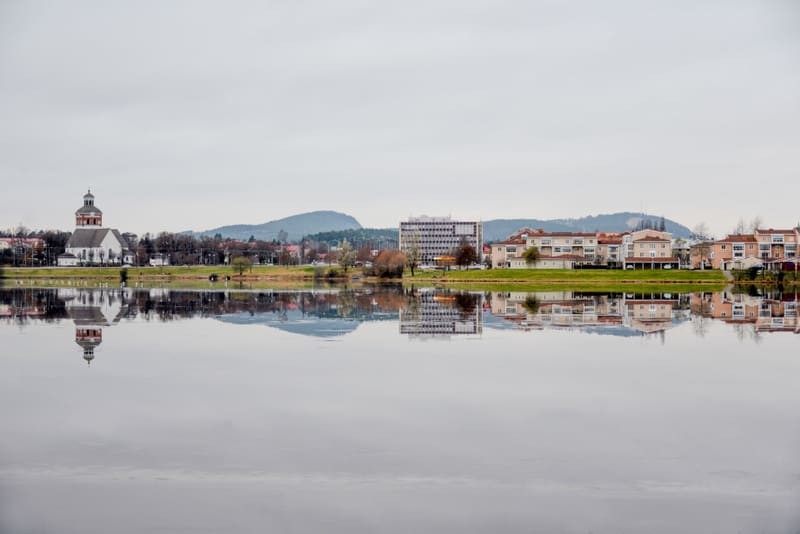 Bilden visar ett landskap med en stad eller tätort som speglar sig i ett stilla vattendrag i förgrunden. På land syns flera byggnader, inklusive en vit kyrka med ett upphöjt klocktor, en stor kontorsliknande byggnad med många fönster, och flera flerf