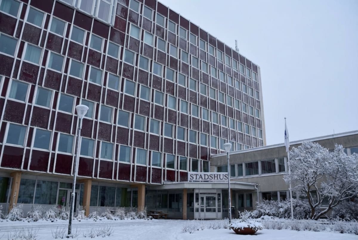 En exteriörbild tagen under dagtid föreställande en stor byggnad med texten "STADSHUS" ovanför entrén. Byggnaden är hög med många fönster och är klädd i ett mörkrött material med vita ramar runt fönstren. Fasadens yta verkar täckas av ett tunt lager snö eller frost, vilket även syns på fönsterkarmarna. Framför entrén finns en snötäckt markyta med buskar och en gatulykta. Bredvid huvudbyggnaden syns en lägre byggnadsdel med ett vitt flaggspel. Ett snöklätt träd och ytterligare buskar syns i förgrunden till höger. Marken är täckt av ett jämnt lager snö. Himmeln är ljusgrå och molnig.