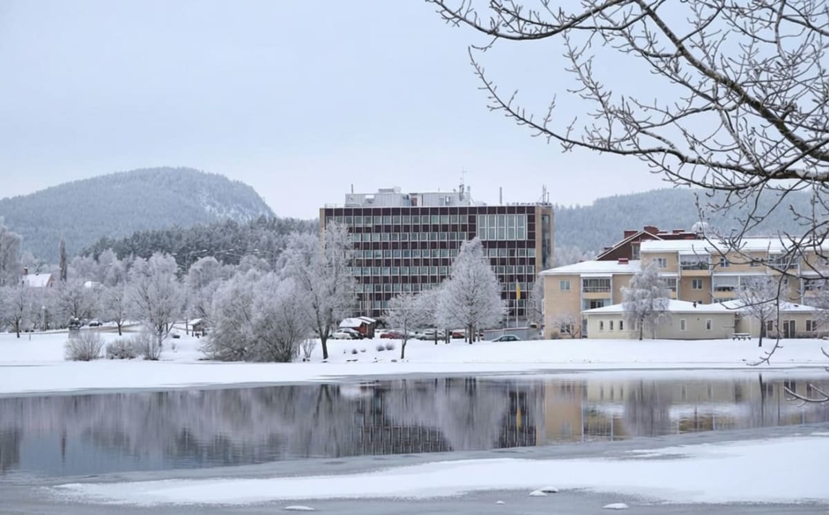 Bilden visar ett vinterlandskap med ett delvis fruset vattendrag i förgrunden, som reflekterar den omgivande miljön. Till vänster om vattendraget finns snötäckta markytor och träd med frostiga grenar. I bakgrunden reser sig en snötäckt bergssluttning med barrskog. Fler träd med frosttäckta grenar syns i mitten av bilden, mellan vattendraget och byggnaderna. En stor, modern byggnad med många fönster och en mörkbrun fasad dominerar mittenpartiet och till höger om den finns en lägre, ljusare byggnad med flera våningar och balkonger. Himlen är ljus och molnig.