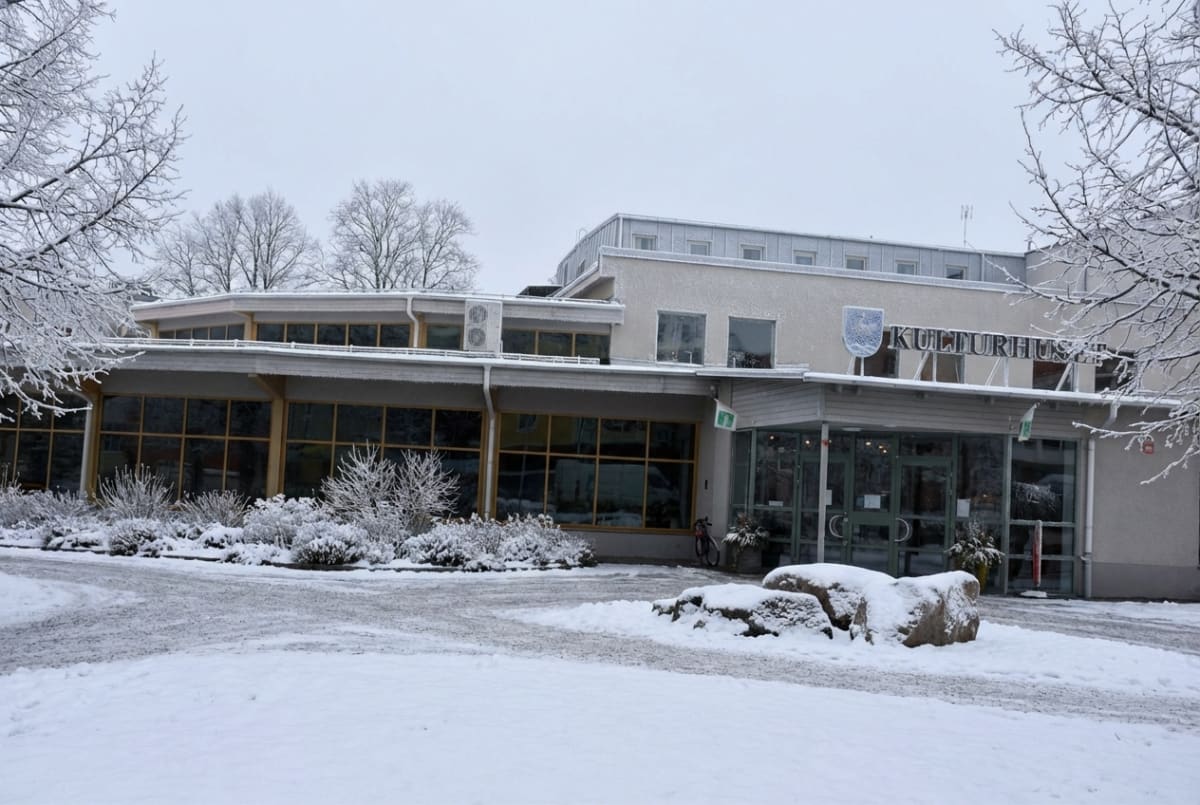 Bilden visar ett kulturhus med delvis snötäckta byggnadspartier och en vintermiljö. Huset har en ljus fasad med stora fönsterpartier och taket är täckt av ett tunt lager snö. På ingångspartiet, som är av glas, syns bokstäverna "KULTURHUS" samt en vapensköld ovanför. Framför byggnaden finns snötäckta buskar och träd, och marken är täckt av ett jämnt snötäcke. En del av gångvägen framför kulturhuset är synlig, delvis snötäckt men med en tydlig gångyta. I förgrunden till höger ligger flera stora stenar, även de delvis snötäckta. Ett par cyklar står parkerade nära entrén. Himlen är grå och mulen, vilket bidrar till vinteratmosfären.