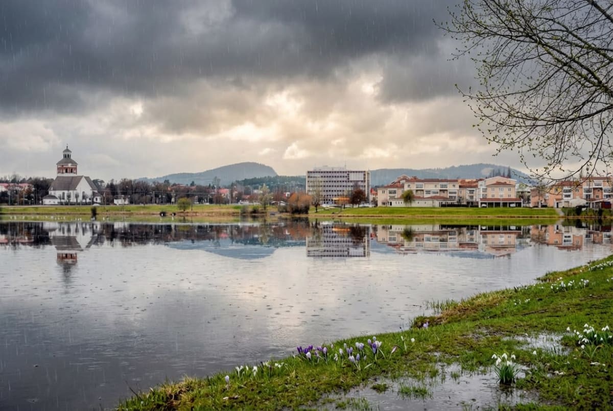 En bild som visar en stadssiluett reflekterad i ett vattendrag under ett regnigt och molnigt väder. I förgrunden till höger syns en gräsmatta med vattenpölar och utslagna vårblommor som snödroppar och krokusar. Vänster om gräsmattan breder ett vattendrag ut sig, där byggnader och himmel speglas tydligt i ytan. På andra sidan vattnet finns en stad med en stor vit kyrka med en rödbrun kupol och spetsig topp till vänster. Flera andra byggnader, inklusive ett höghus, syns också längs strandkanten. Bakom staden höjer sig kullar och berg mot horisonten. Himmelen är täckt av mörka regnmoln, och regnstreck syns tydligt i luften, särskilt i överkant av bilden. Ett ensamt träd med spirande lövverk syns i bildens övre högra hörn.