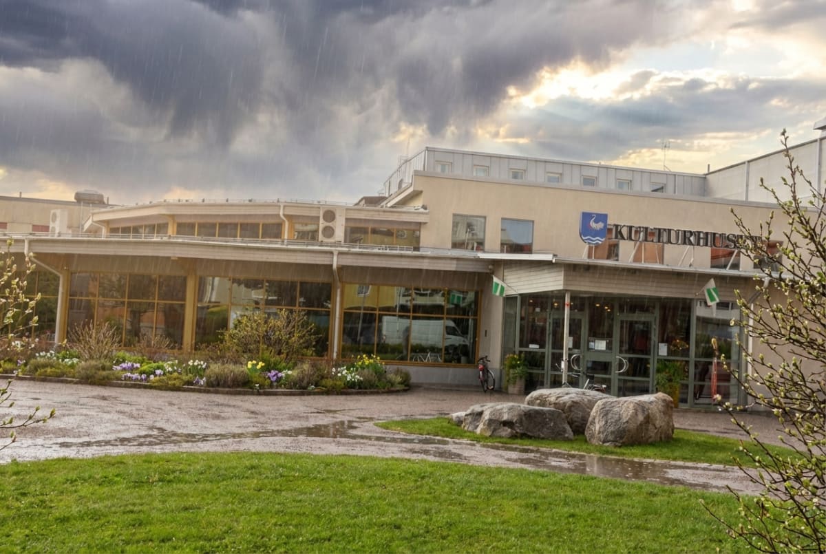 En exteriörbild tagen under dagtid föreställande en byggnad märkt "KULTURHUSET" mot en molnig himmel med synligt regn. Byggnaden har en ljusgul fasad och stora fönsterpartier. Framför byggnaden finns en gräsyta, en asfalterad gångväg med pölar av vatten, samt en rabatt med vårblommor i lila, gult och vitt. Några stora stenblock är placerade vid gångvägen. Gröna flaggor med vita kors och en cykel syns nära byggnadens entré. En logotyp med en svan eller fågel i blått och vitt sitter ovanför ordet "KULTURHUSET". Regn faller synligt över hela bilden, och himlen är täckt av tunga, grå moln, men med en ljusare del mot höger och bakom byggnaden.