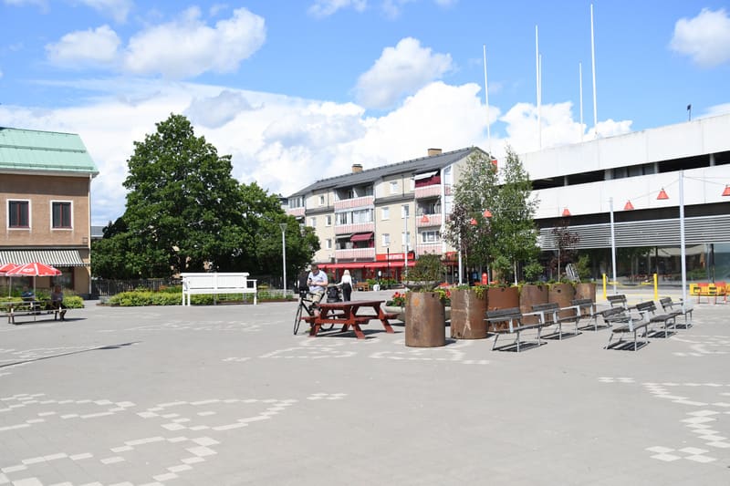 Bilden visar ett torg eller en öppen plats utomhus under dagtid. Torget är belagt med ljusa stenplattor i ett mönster. Till vänster i bilden syns en byggnad med en tegelfärgad fasad, grönt tak och ett röd- och vitrandigt markis. Framför den byggnaden