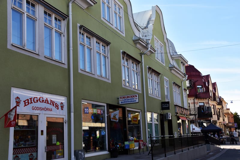 Bilden visar fasaden på flera byggnader längs en gata med en klarblå himmel i bakgrunden. I förgrunden till vänster syns en byggnad med en matt grön fasad och vita fönsterkarmar. På bottenvåningen finns två butiker: "BIGGANS GODISHÖRNA" med en rödvit