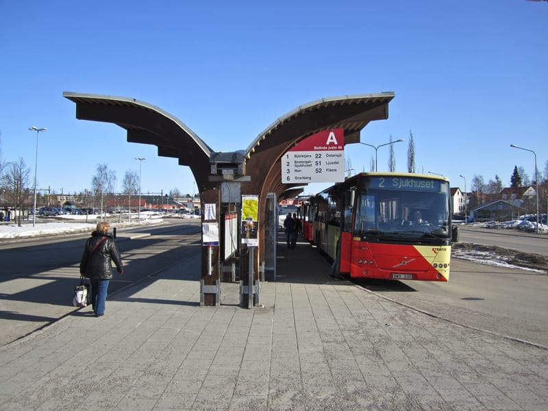 En bild tagen utomhus under dagtid som visar en busshållplats och en buss. En person med mörk jacka och ljus väska går bort från kameran längs en gångväg vid busshållplatsen. Hållplatsen har ett överdrag i trä och metall samt informationsskyltar med