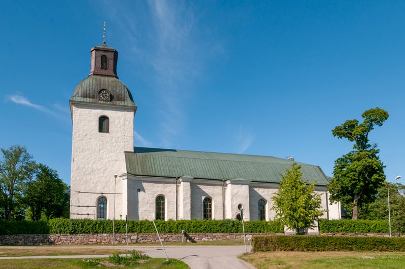 Bilden visar en medeltida kyrka med vit putsad fasad och ett grönt tak. Kyrkan har en stor kvadratisk torn med en kopparklädd hjälm och en spira med ett kors på toppen. Högst upp på tornet syns en klocka. Kyrkans långhus har flera välvda fönster och
