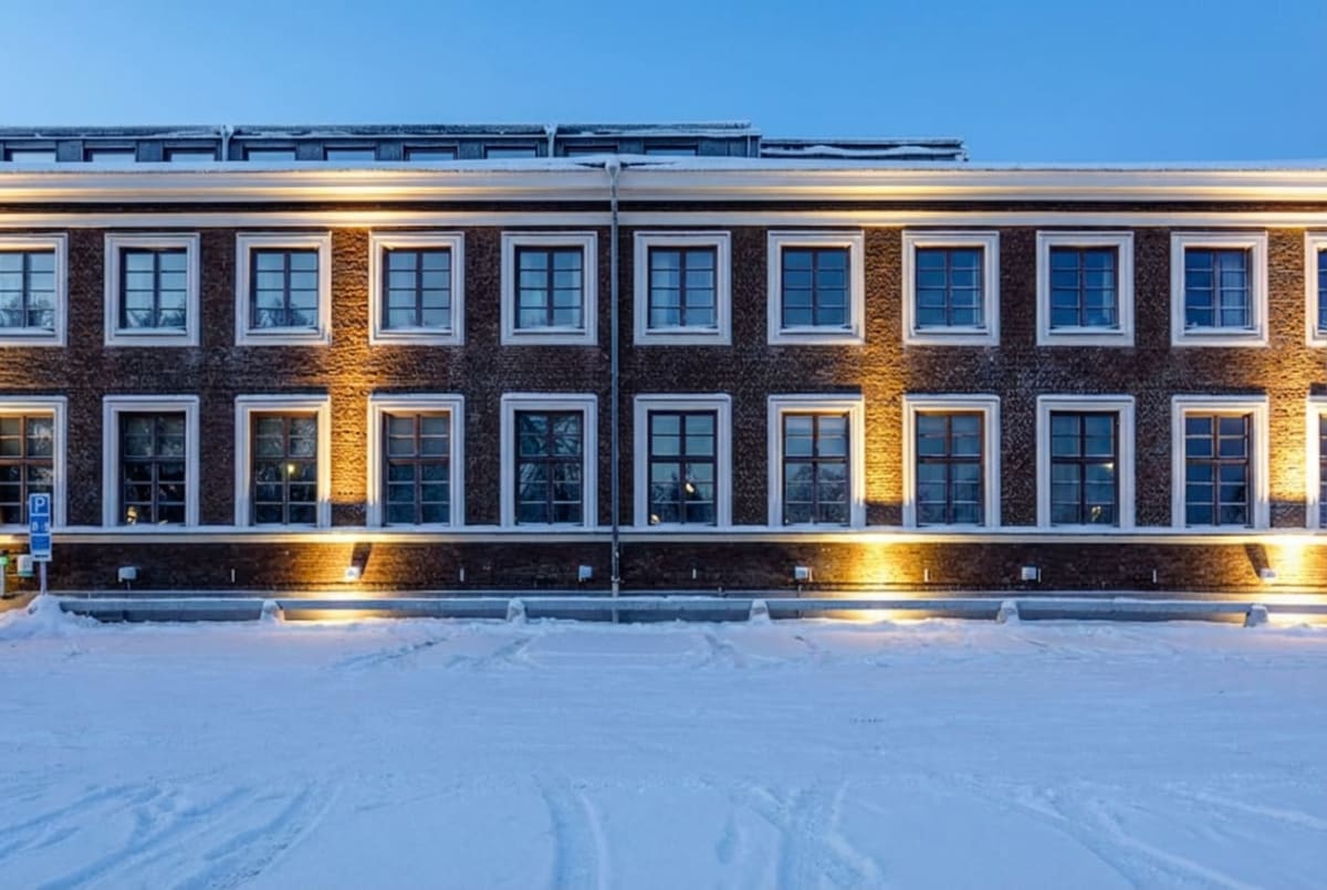 Bilden visar fasaden på en byggnad under vintertid. Byggnaden är upplyst med varmt ljus från belysning monterad under fönstren, vilket skapar kontraster mot den snötäckta marken framför. Marken är täckt av ett tjockt lager snö med spår av fordon. Fasaden är av tegel och har flera fönsterrader med vita ramar och spröjsar. Ovanför nedersta fönsterraden sträcker sig en horisontell vit list. Taket, som syns delvis, är också täckt av snö. En blå parkeringsskylt med ett 'P' och texten '8-18' är synlig till vänster framför byggnaden. Himlen är klar och blå. Ljusskenet från lamporna reflekteras i den vita snön.