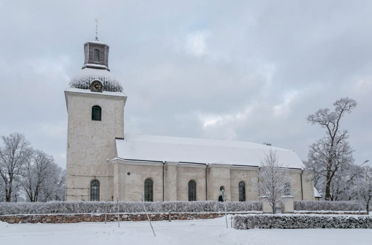 En vit kyrkobyggnad med ett högt torn är centralt placerad i bilden. Kyrktornet har en svart klocka med gyllene visare och siffror, samt en spira på toppen. Byggnadens tak och torn är täckta med ett lager snö. Runt kyrkan finns snötäckta buskar och träd, varav vissa träd är snöbeklädda och utan löv. Ett staket av sten syns i förgrunden, som avgränsar kyrkan från ett snötäckt markområde. Himmelen är mulen och grå.