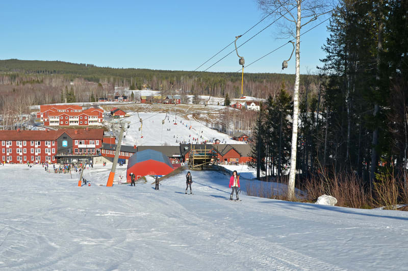 En vidvinkelbild tagen på en solig vinterdag som visar en skidbacke full med snö i förgrunden och mellangrunden. I bakgrunden syns skogsklädda berg. Flera personer, både vuxna och barn, är klädda i vinterkläder och åker skidor eller snowboard nedför