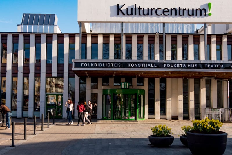 Bilden visar fasaden till en större byggnad med texten "Kulturcentrum!" på taket. Nedanför taket syns en markis med texten "FOLKBIBLIOTEK KONSTHALL FOLKETS HUS TEATER". Byggnaden har vertikala fält med rutor av fönster och mörkare paneler. Framför by