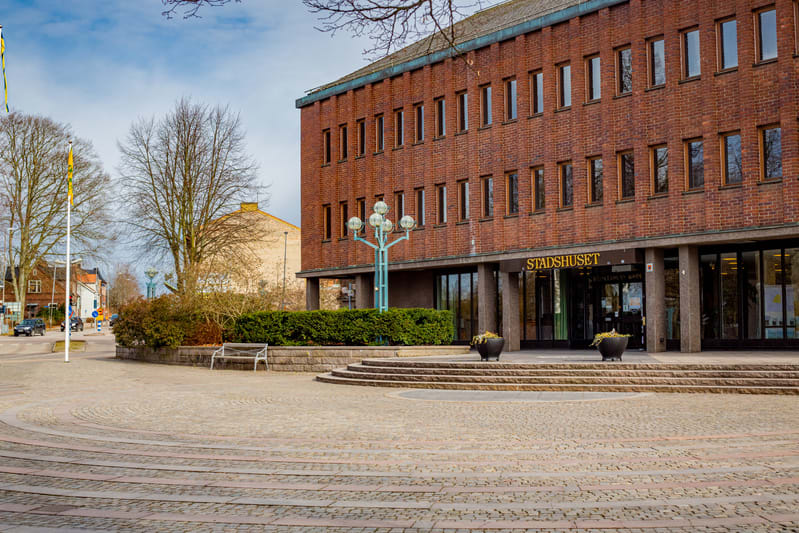 Bilden visar fasaden till en större tegelbyggnad med texten "STADSHUSET" ovanför huvudentrén. Ingången består av flera glasdörrar och fönster. Framför byggnaden finns ett stenlagt torg med cirkulära mönster och en trappa som leder upp mot entrén. Til