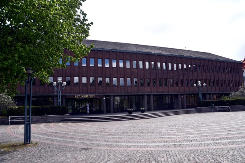En bild tagen utomhus visar en tegelbyggnad med flera rader av fönster och en platt taklinje. Byggnaden har skylten "STADSHUSET" ovanför entrén. Framför byggnaden finns en stor öppen yta belagd med kullersten i ett cirkulärt mönster. En trappa leder
