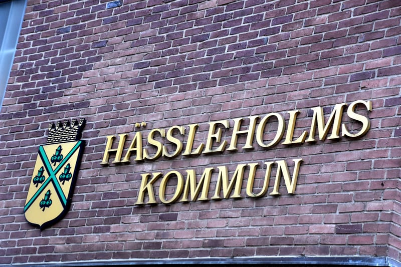 En bild som visar en skylt på en brun tegelvägg. Skylten, som är i guldglänsande bokstäver, stavar "HÄSSLEHOLMS KOMMUN". Till vänster om texten finns en vapensköld. Vapenskölden har en gulorange bakgrund med ett grönt kryss. I varje kvadrat bildad av