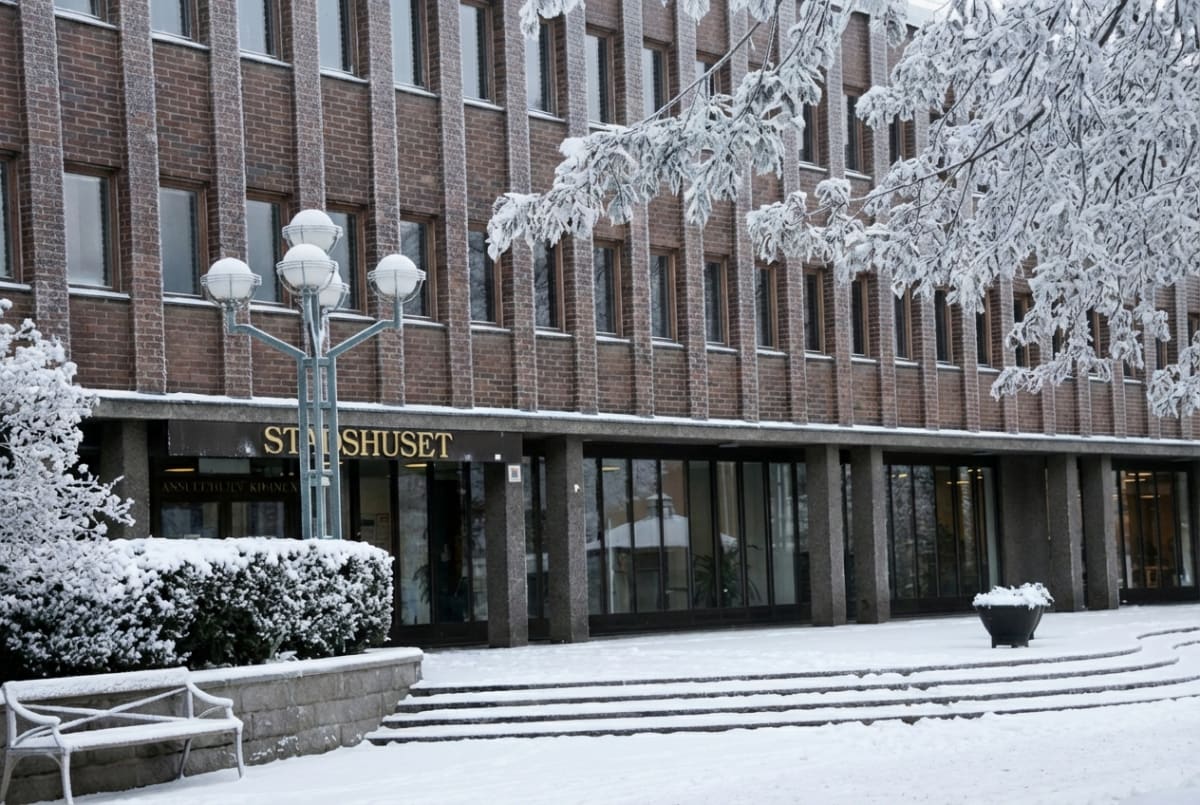 Bilden visar fasaden på en byggnad under vintertid, täckt med snö. Byggnaden har en tegelfasad med vertikala betongdetaljer och flera rektangulära fönster. Entrén på bottenvåningen består av stora glaspartier. Till vänster om entrén finns en buske och ett träd, båda täckta av snö. En vit parkbänk står framför busken. Framför entrén leder breda trappsteg upp till byggnaden, även dessa täckta av snö. Ett flerstammigt träd med snötyngda grenar hänger in från höger, och i förgrunden finns en gatlykta med flera klotformade lampor, också snötäckt. Ovanför entrén står texten "STADSHUSET" i guldiga bokstäver.