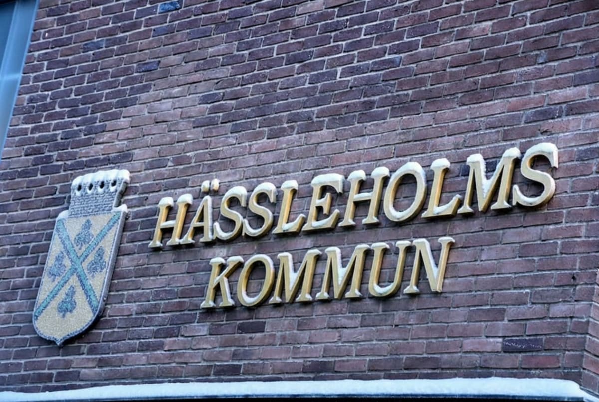 Bilden visar fasaden på en byggnad, tillverkad av rött tegel. På fasaden sitter texten "HÄSSLEHOLMS KOMMUN" i upphöjda guldfärgade bokstäver. Övanpå bokstäverna ligger ett tunt lager av snö eller is. Till vänster om texten finns en vapensköld som också har ett tunt lager snö på sig. Vapenskölden är dekorerad med ett mönster i blått, grönt och gult, och är krönt med en murkrona. Längst ner på bilden anas en vit yta, eventuellt en snötäckt yta eller en del av byggnadens grund.