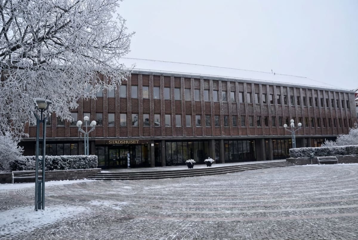 Bilden visar ett stort Stadhus i tegel med en snötäckt framsida på en vinterdag. Byggnaden har många fönster i flera rader och en tydlig skylt med texten ”STADHUSET” över huvudentrén. Framför byggnaden finns ett stenlagt torg som delvis är täckt av snö. Till vänster i bilden syns grenarna på ett träd som är täckta av snö eller frost. Det finns även snöbeklädda buskar framför byggnaden och gatlyktor på torget. Hela scenen ger ett intryck av en kall vinterdag.